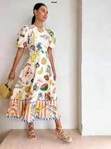 Woman wearing a white midi dress with a colorful tropical fruit and bird print, puff sleeves, and a striped hem with seafood motifs, holding a yellow handbag and standing against a white textured wall.