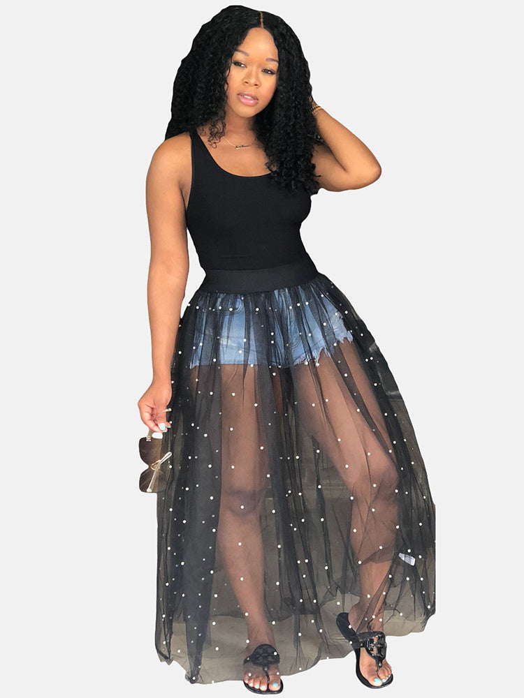 Woman holding sunglasses, wearing a black tank top and a black sheer maxi skirt with white pearl embellishments over denim shorts.