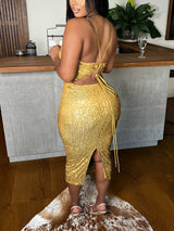 Woman wearing a gold sequined midi dress with spaghetti straps and a backless design with lace-up detail, back view