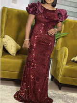 Woman wearing a long, shimmering burgundy sequined dress with ruffled cap sleeves, standing between two gold chairs.