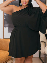 Close-up of a plus-size woman smiling, wearing a black one-shoulder dress with an asymmetrical sleeve and braided hair.