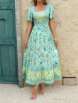 Woman in a light green floral print midi dress with a square neckline, leaning against a wall.