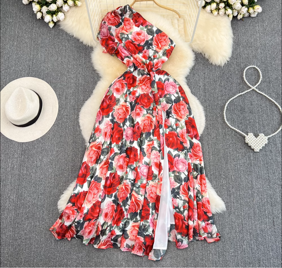 A floral print one-shoulder maxi dress with a thigh-high slit, laid flat on a grey rug with a white hat and pearl necklace.