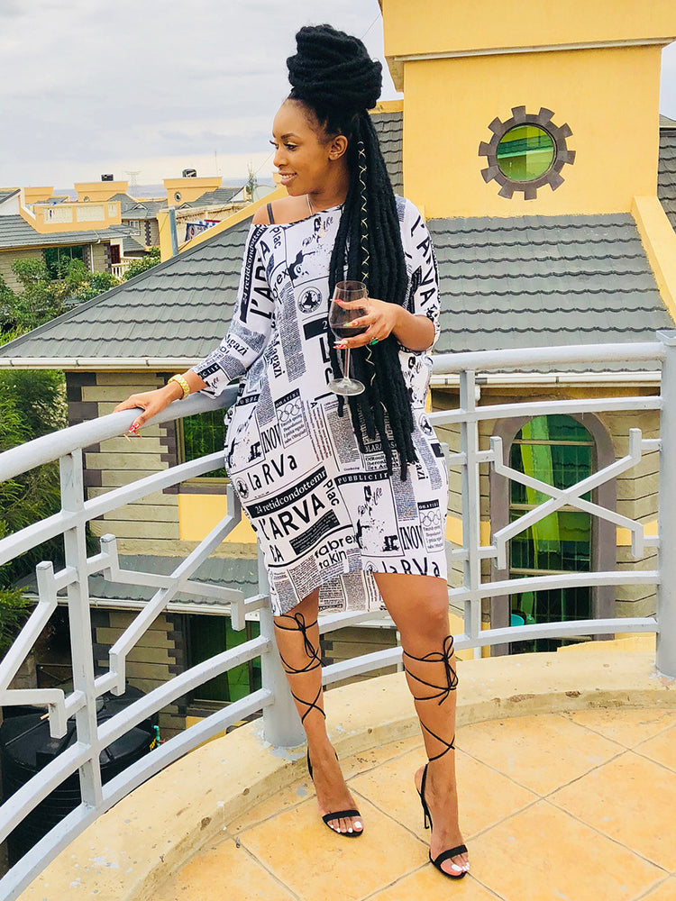 Woman wearing a black and white newspaper print t-shirt dress with black strappy heels, standing on a balcony.