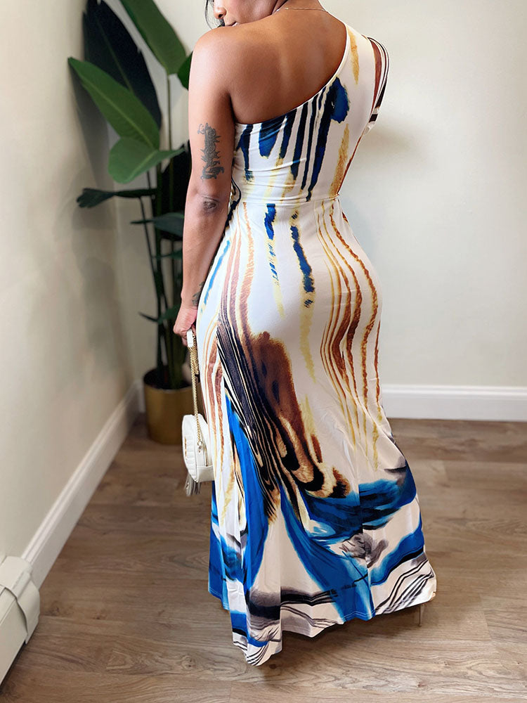 Back view of a woman wearing a one-shoulder maxi dress with an abstract print, holding a white handbag.