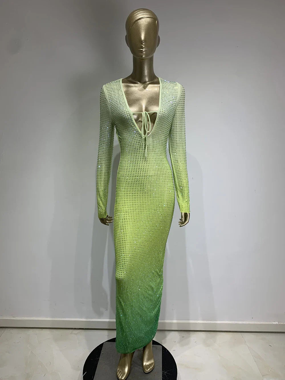 A mannequin displays a long, gradient green dress with long sleeves and a plunging neckline, embellished with rhinestones.