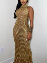 Woman wearing a long, gold beaded, sheer halter neck gown, posing against a white wall.