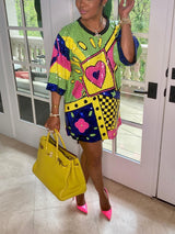 Side view of a woman in a vibrant sequined t-shirt dress with geometric and graphic details, accessorized with a yellow handbag and pink heels.