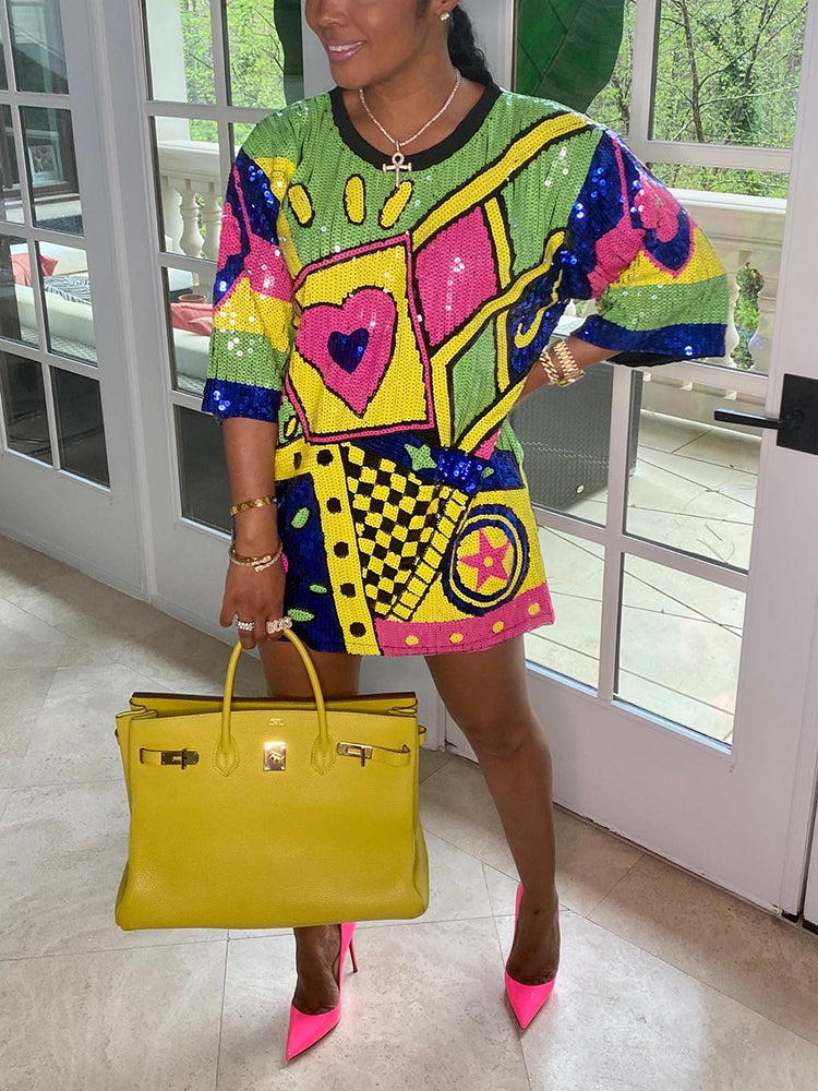 Woman wearing a colorful sequined t-shirt dress with a heart graphic and geometric patterns, holding a yellow handbag and wearing pink heels.