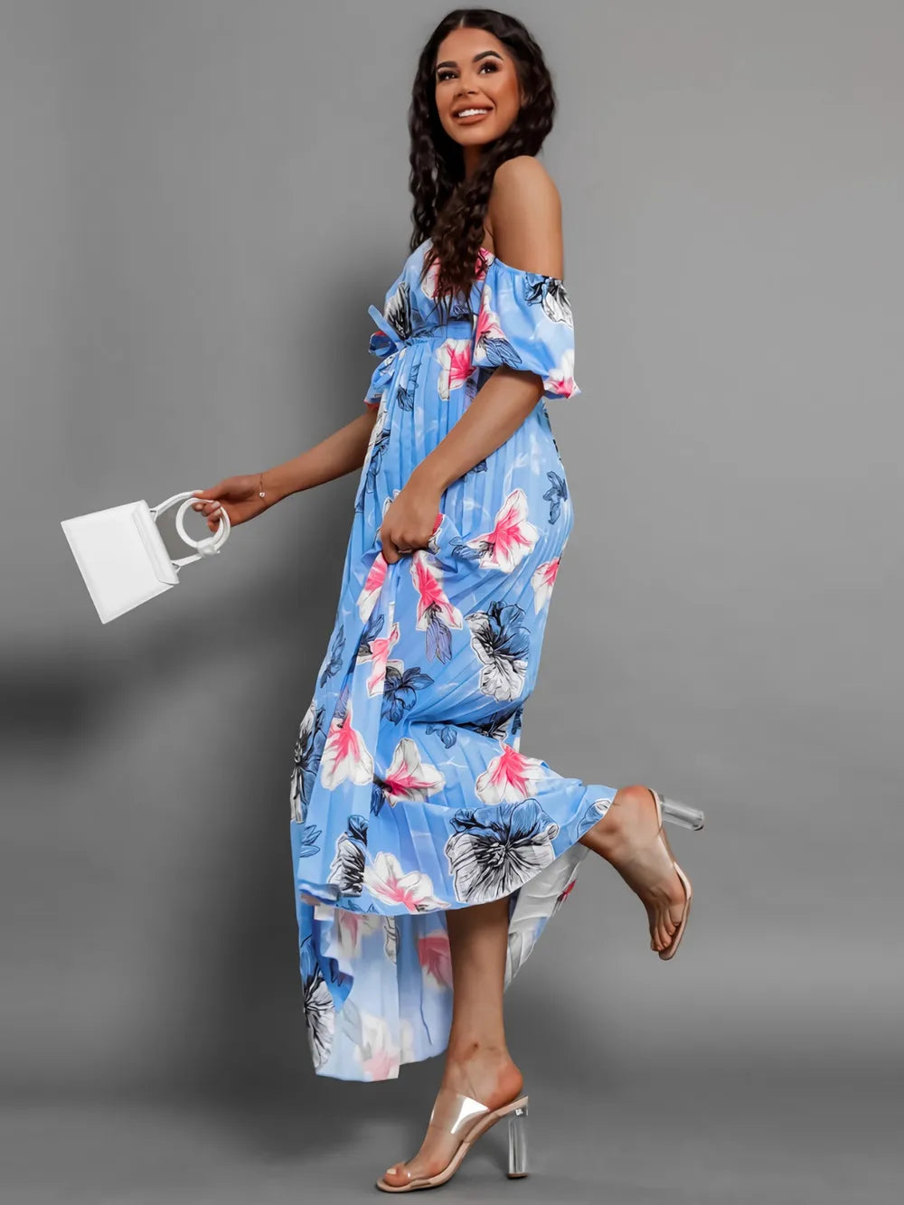 Woman in a blue floral maxi dress with off-the-shoulder neckline and pleats, walking with a white handbag.