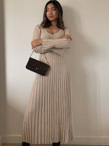 Woman with dark hair wearing a beige ribbed knit maxi dress with flared cuffs and a brown monogrammed handbag.