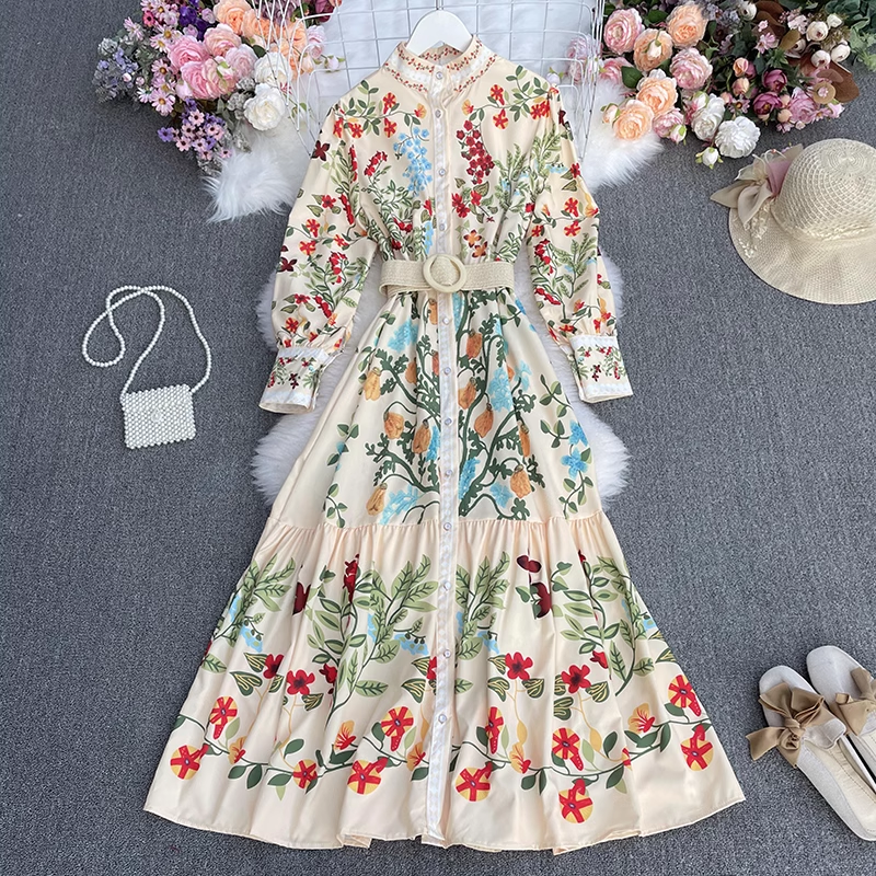 Cream floral print maxi dress with long sleeves and a belted waist, laid flat on a grey background with decorative flowers and accessories.