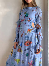 Side front view of a woman in a light blue graphic print maxi dress with cut-out waist detail and long, flared sleeves.