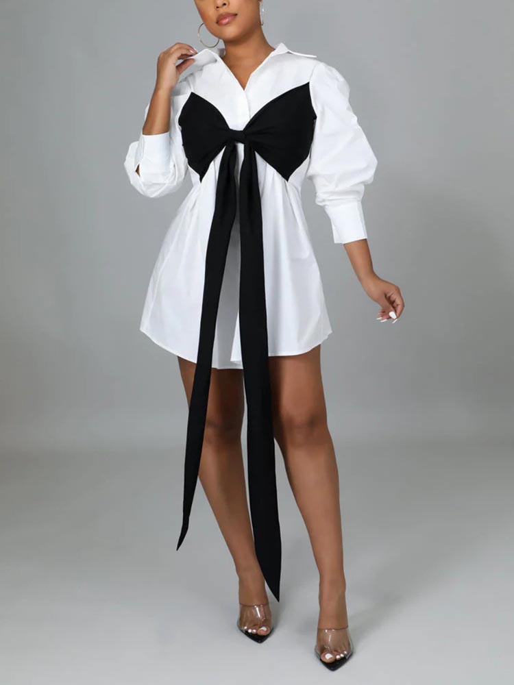 Model wearing a short white shirt dress with a black bow, paired with clear heels.