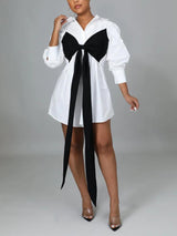 Model wearing a short white shirt dress with a black bow, paired with clear heels.