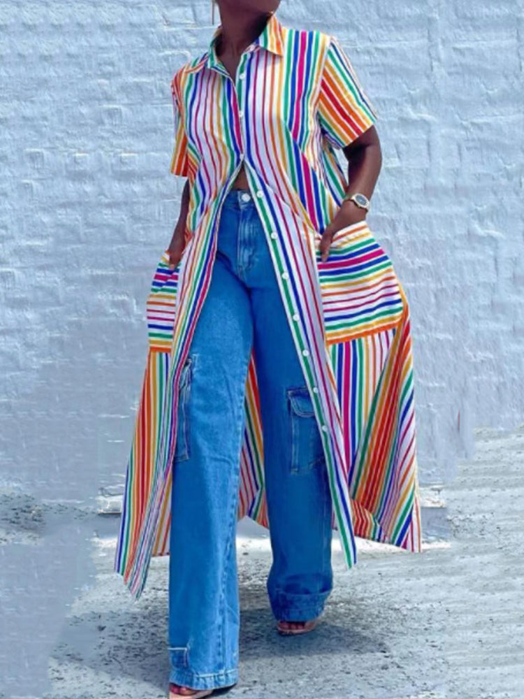 Woman wearing a long rainbow striped shirt dress unbuttoned over blue wide-leg jeans, showing front pockets, standing against a white brick wall.