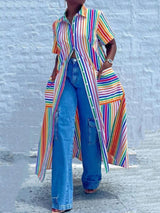 Woman wearing a long rainbow striped shirt dress unbuttoned over blue wide-leg jeans, showing front pockets, standing against a white brick wall.