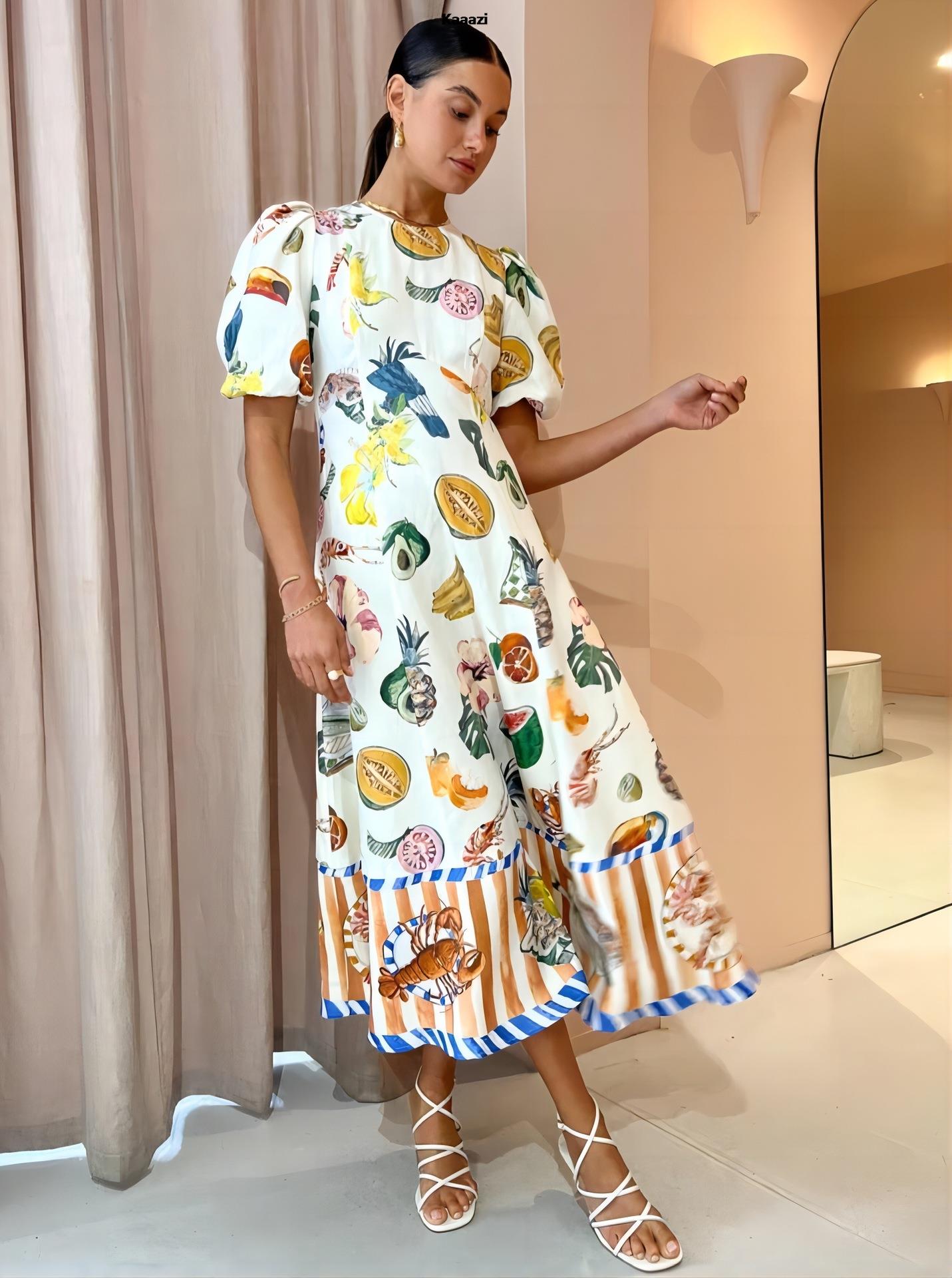 Woman wearing a white midi dress with a colorful tropical fruit and bird print, puff sleeves, and a striped hem with seafood motifs, standing indoors with a curtain in the background.