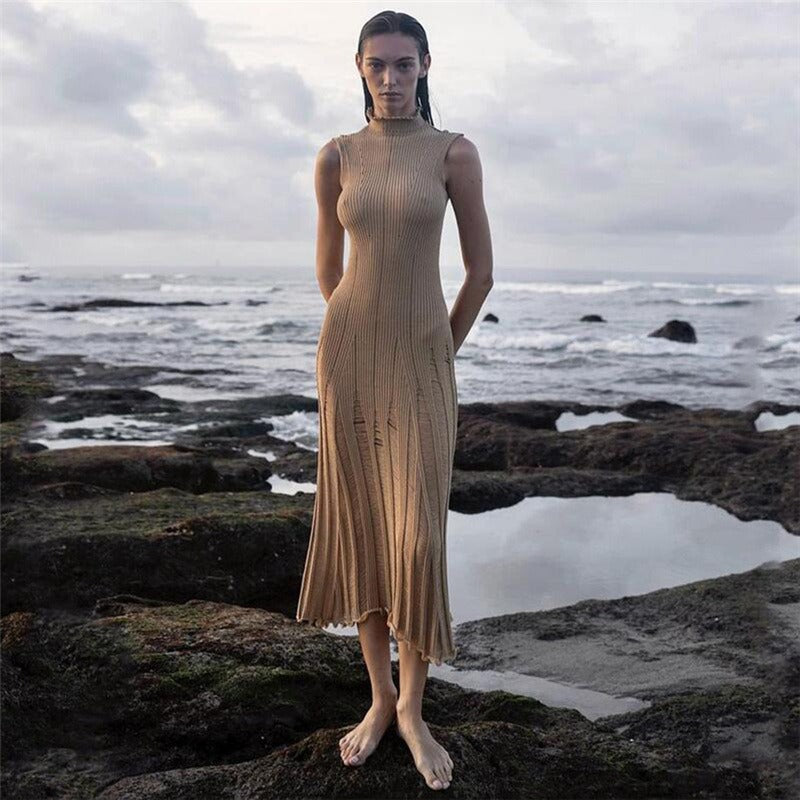 Woman wearing a beige sleeveless ribbed knit midi dress with sheer panel detailing standing on rocks by the ocean