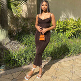 Woman wearing a brown sleeveless midi dress with spaghetti straps and a cowl neckline, accessorized with a small orange handbag and white strappy heels, standing outdoors.
