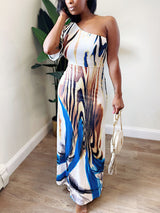 Woman wearing a one-shoulder maxi dress with an abstract print, holding a white handbag.