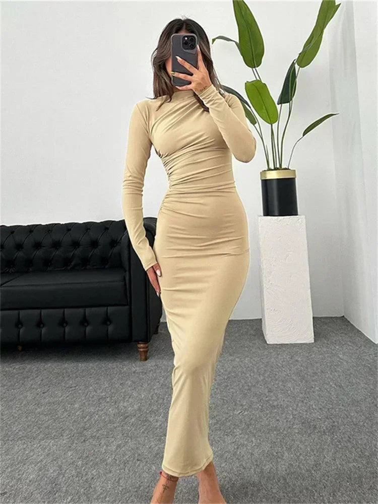Woman wearing a long-sleeved, ruched, beige maxi dress, standing in a room with a black sofa and a plant.