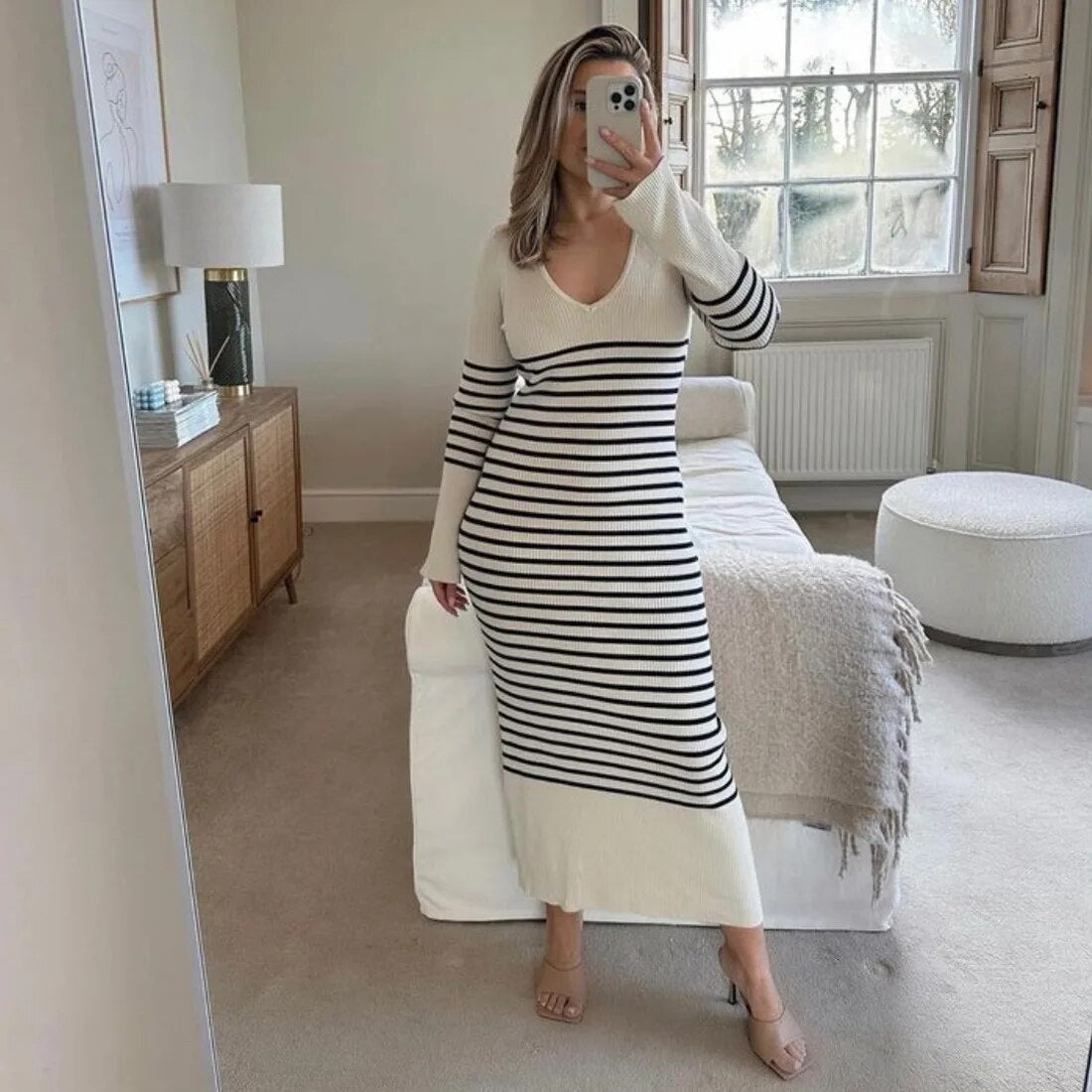 Woman wearing a long-sleeved striped maxi dress and nude heels, taking a mirror selfie in a bedroom
