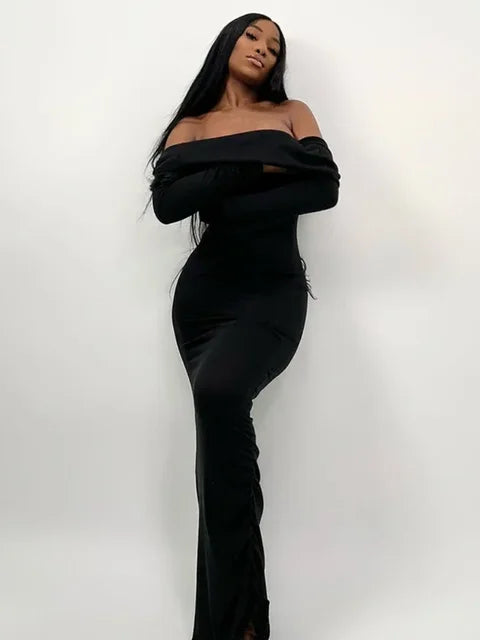 Woman wearing a long black off-the-shoulder dress with long sleeves, leaning against a white wall with arms crossed.