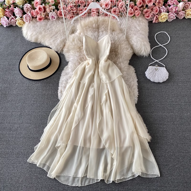 A flowing, cream-colored chiffon dress with ruffle detailing and spaghetti straps, laid flat on a faux fur rug with a straw hat and a beaded clutch.