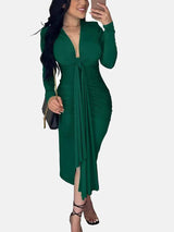 Woman wearing a long-sleeved green ruched dress with a tie-front detail and a slit hem, holding a black purse.