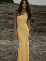 Woman wearing a yellow one-shoulder ribbed knit maxi dress with a mermaid hem, standing on a rocky shore.