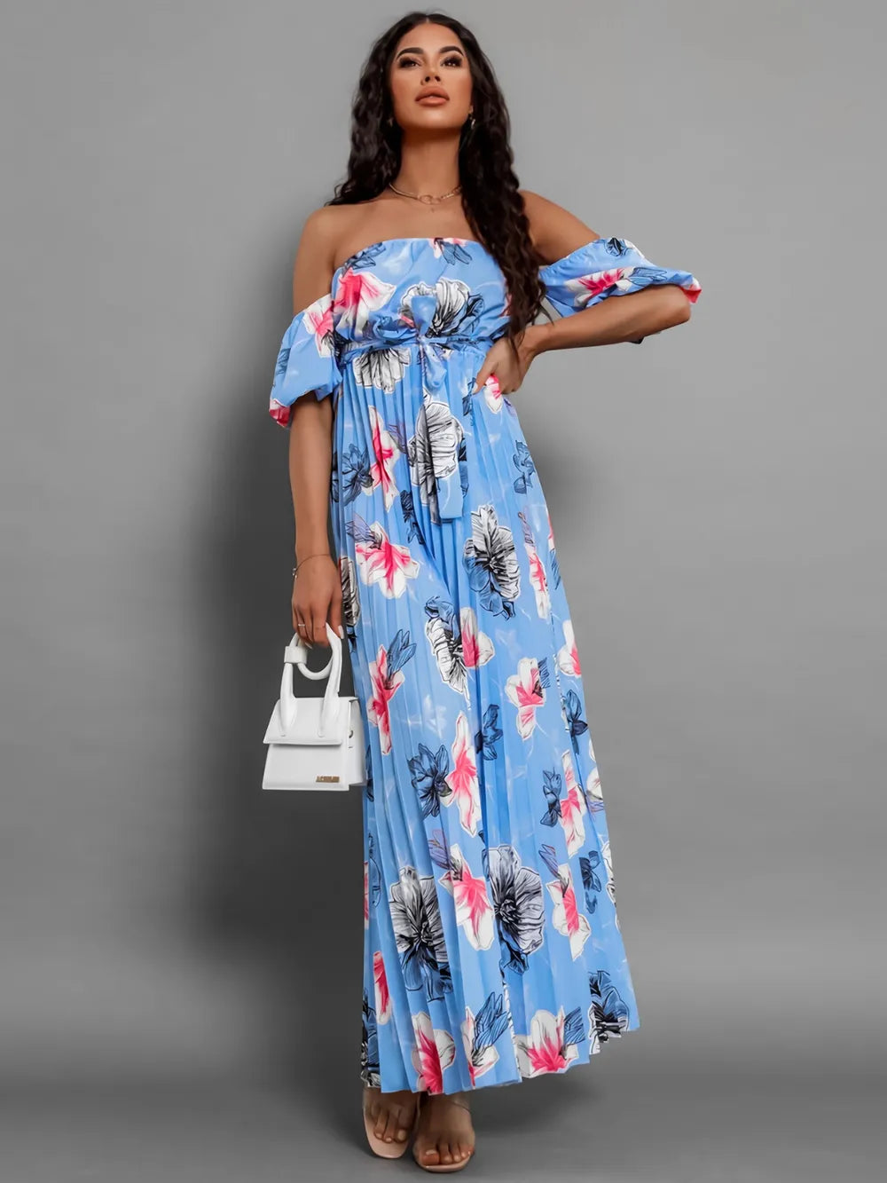 Woman wearing a blue floral print off-the-shoulder maxi dress with short puff sleeves, cinched waist, and pleated skirt, holding a white handbag.