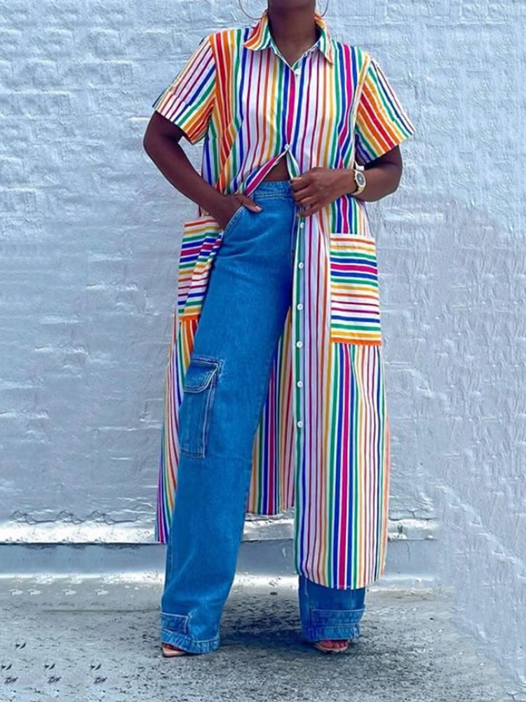 Woman wearing a long rainbow striped shirt dress over blue wide-leg jeans, standing against a white brick wall.
