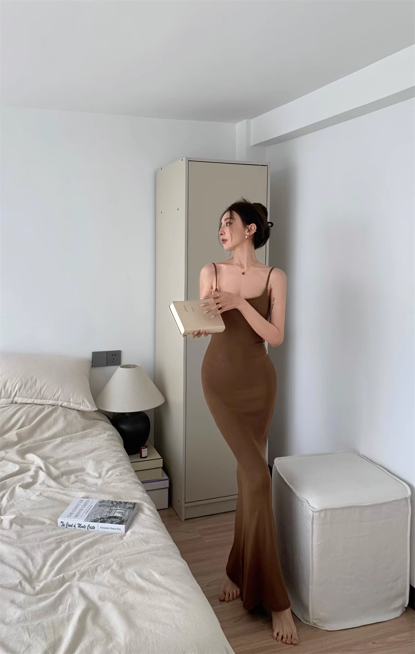 Woman in a brown maxi dress with spaghetti straps holding a book in a bedroom.