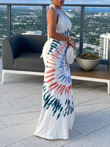 Full body shot of a woman in a white maxi dress with a tie-dye pattern, standing outdoors.