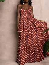 Woman wearing a brown and white tie-dye print maxi dress with a belted waist, adjustable spaghetti straps, and a ruffled hem, holding the skirt slightly.