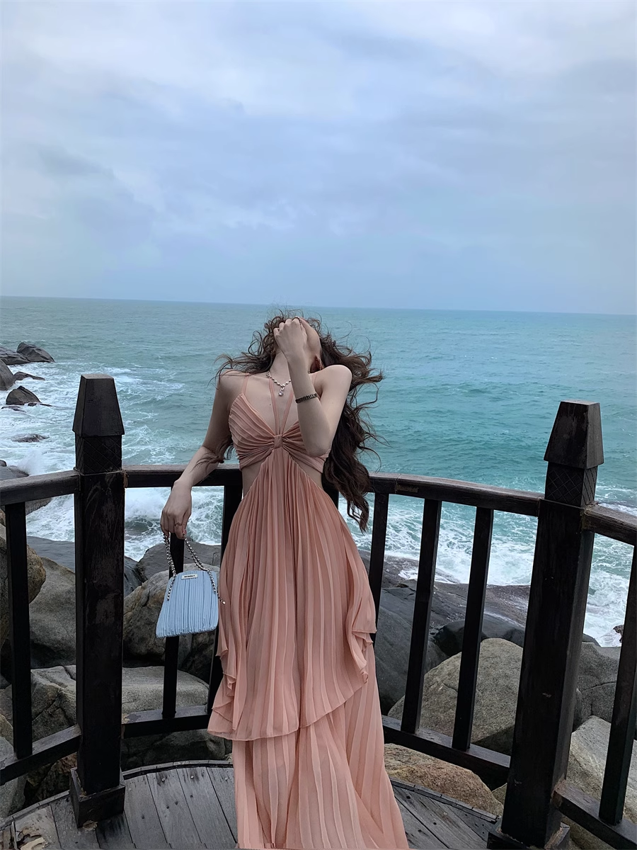 Woman wearing a peach halter neck maxi dress with a cut-out and pleated tiered skirt, standing on a wooden balcony overlooking the ocean.