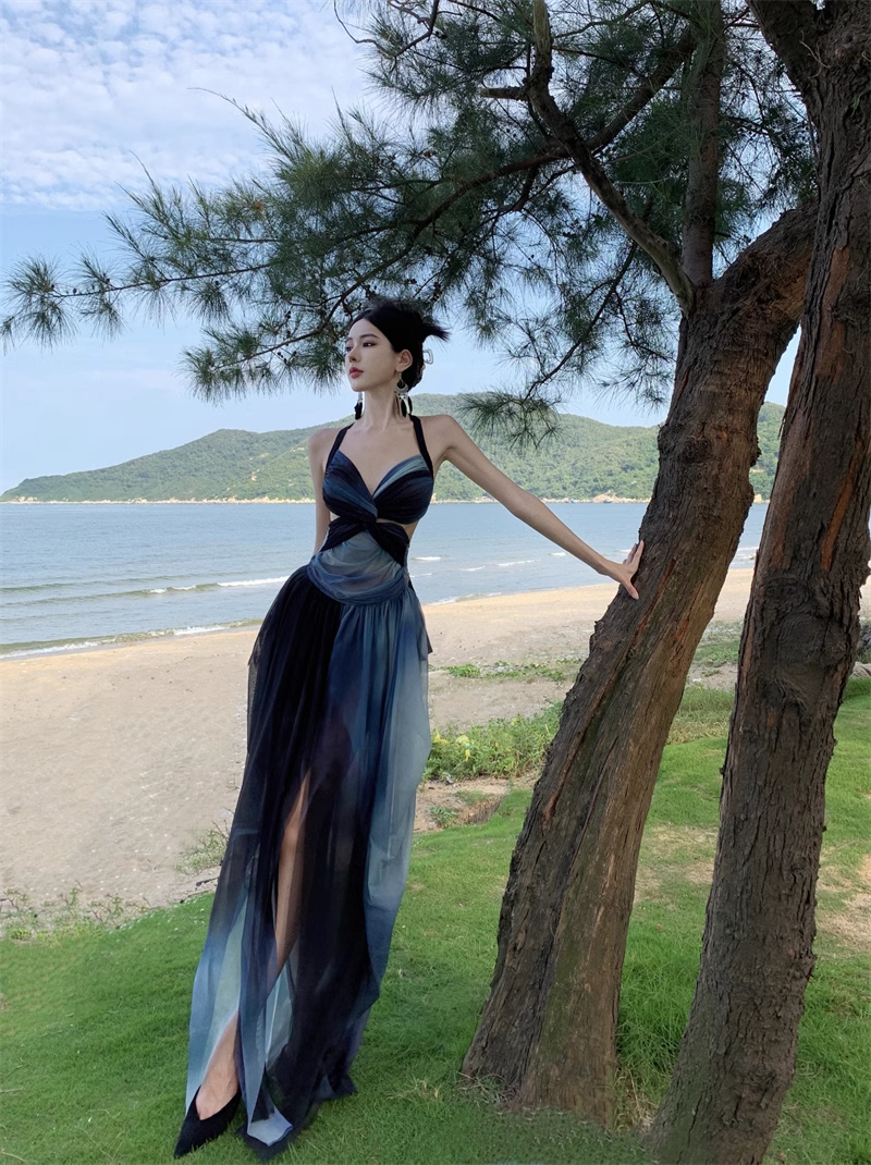 Woman wearing a long, flowing ombre dress with a halter neckline and cutout waist, standing on a grassy area near a beach.
