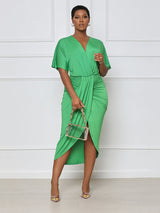 Woman wearing a green V-neck, short-sleeve, ruched, draped midi dress with a tulip hem, holding a clear handbag.