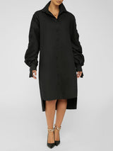 Full front view of a woman wearing a black collared shirt dress with long ruffled sleeves and an asymmetrical hemline, wearing black pointed-toe heels with gold chain accents.