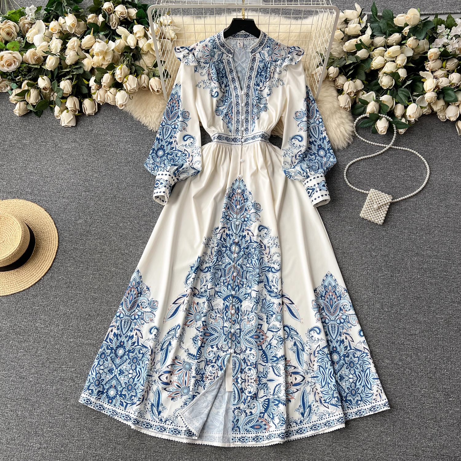 A white maxi dress with a blue paisley print laid flat on a grey surface, adorned with floral arrangements and accessories.