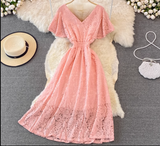 Pink lace dress with V-neck and flutter sleeves displayed on a white fluffy rug, with a straw hat and a white beaded purse nearby.