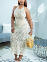 Woman wearing a cream-colored open-knit halter maxi dress with geometric patterns, holding a small woven bag.