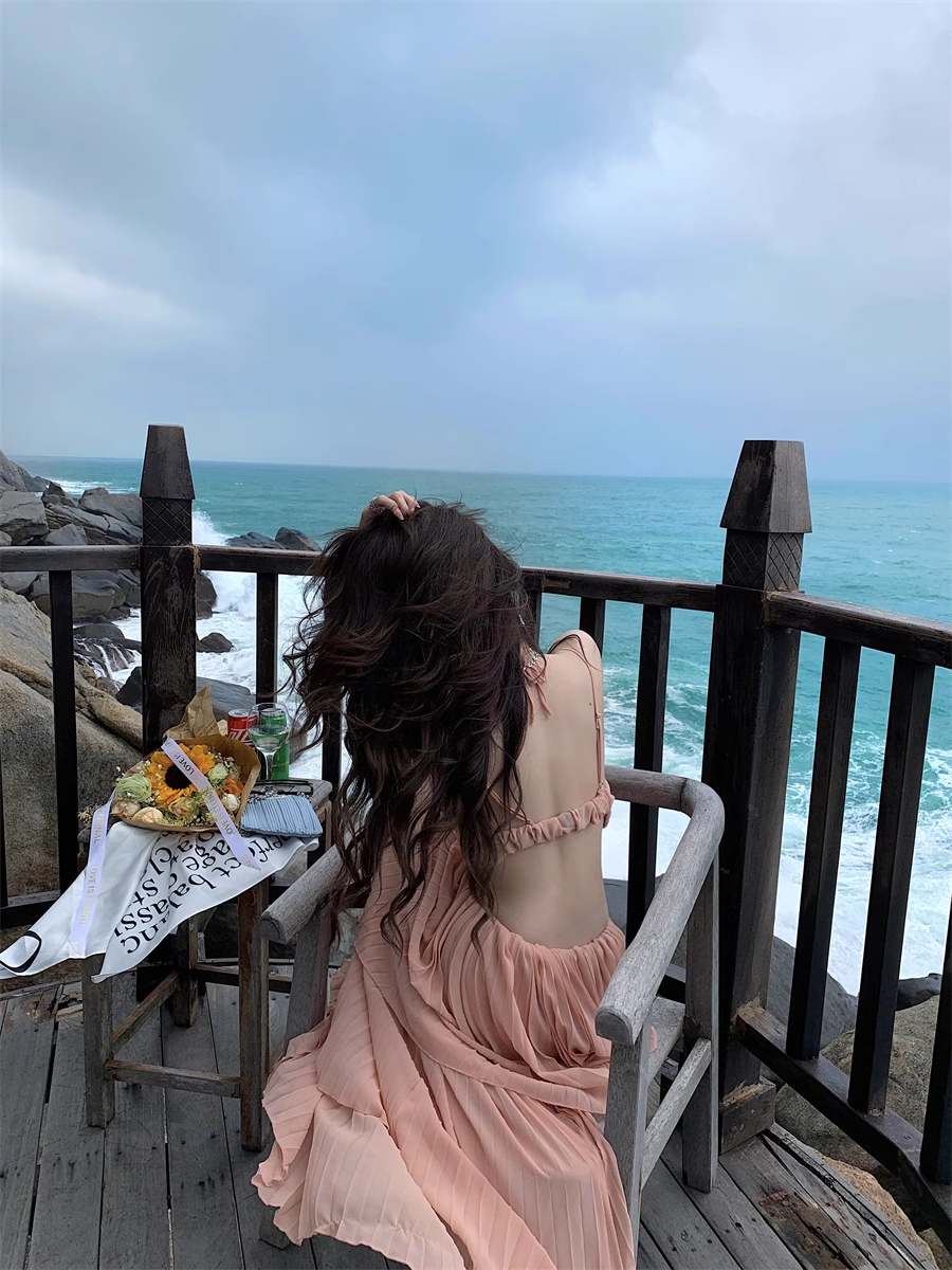 Woman wearing a peach halter neck maxi dress with a cut-out and pleated tiered skirt, sitting on a chair and looking out at the ocean.