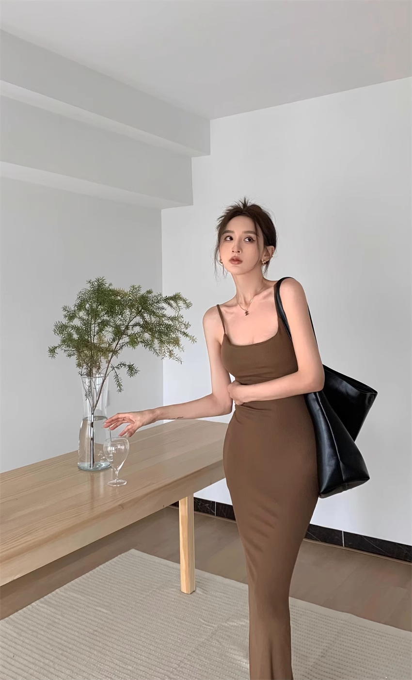 Woman in a brown maxi dress with spaghetti straps standing next to a wooden table with a plant.