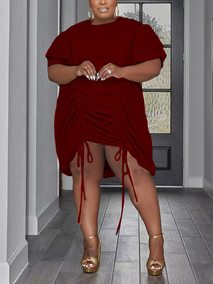 A woman stands in a hallway wearing a burgundy ruched dress with drawstring details and gold platform heels.