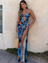 Woman wearing a blue abstract print strapless maxi dress with cut-outs and a high slit, holding a straw bag.