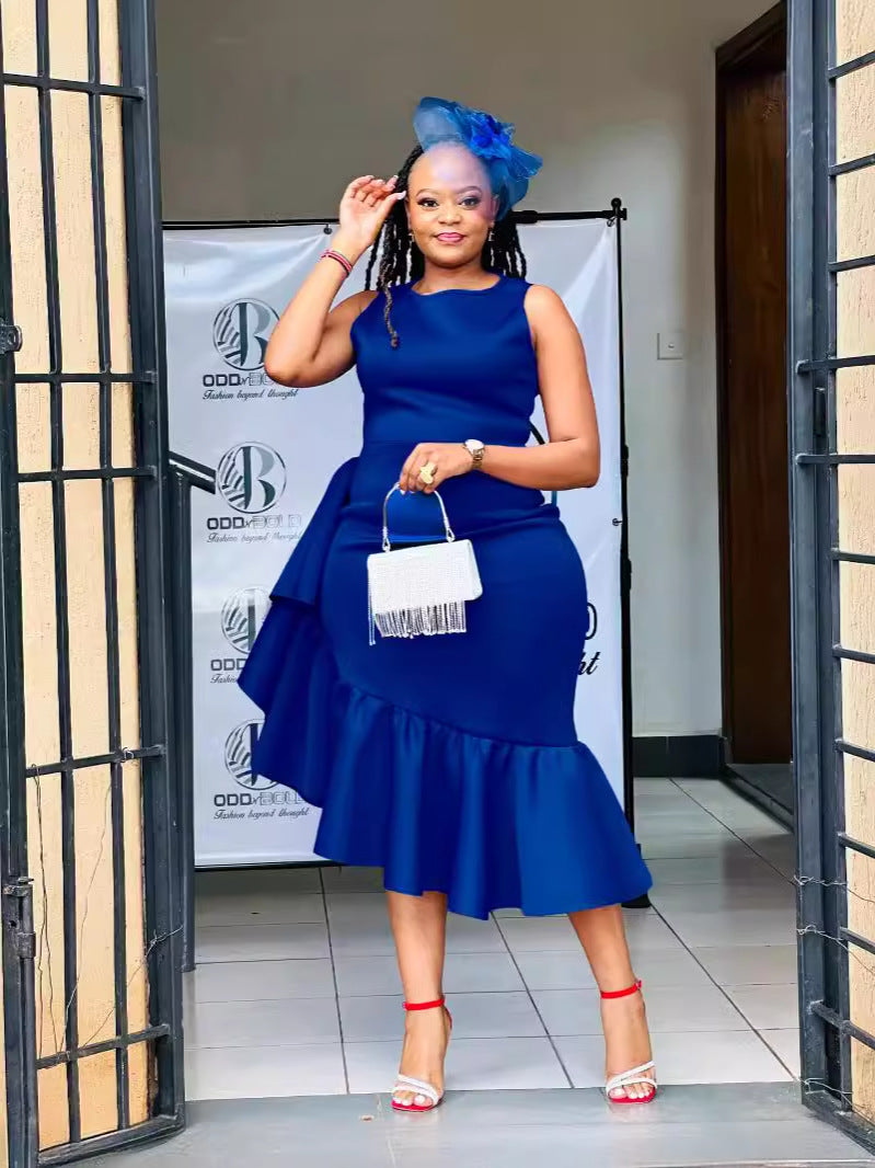 Woman wearing a royal blue sleeveless dress with ruffle detailing and an asymmetrical hemline, accessorized with a blue fascinator and silver handbag.