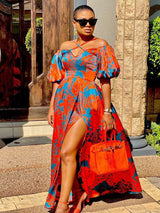 Woman wearing a vibrant orange and blue floral print off-the-shoulder maxi dress with a high leg slit and puff sleeves.