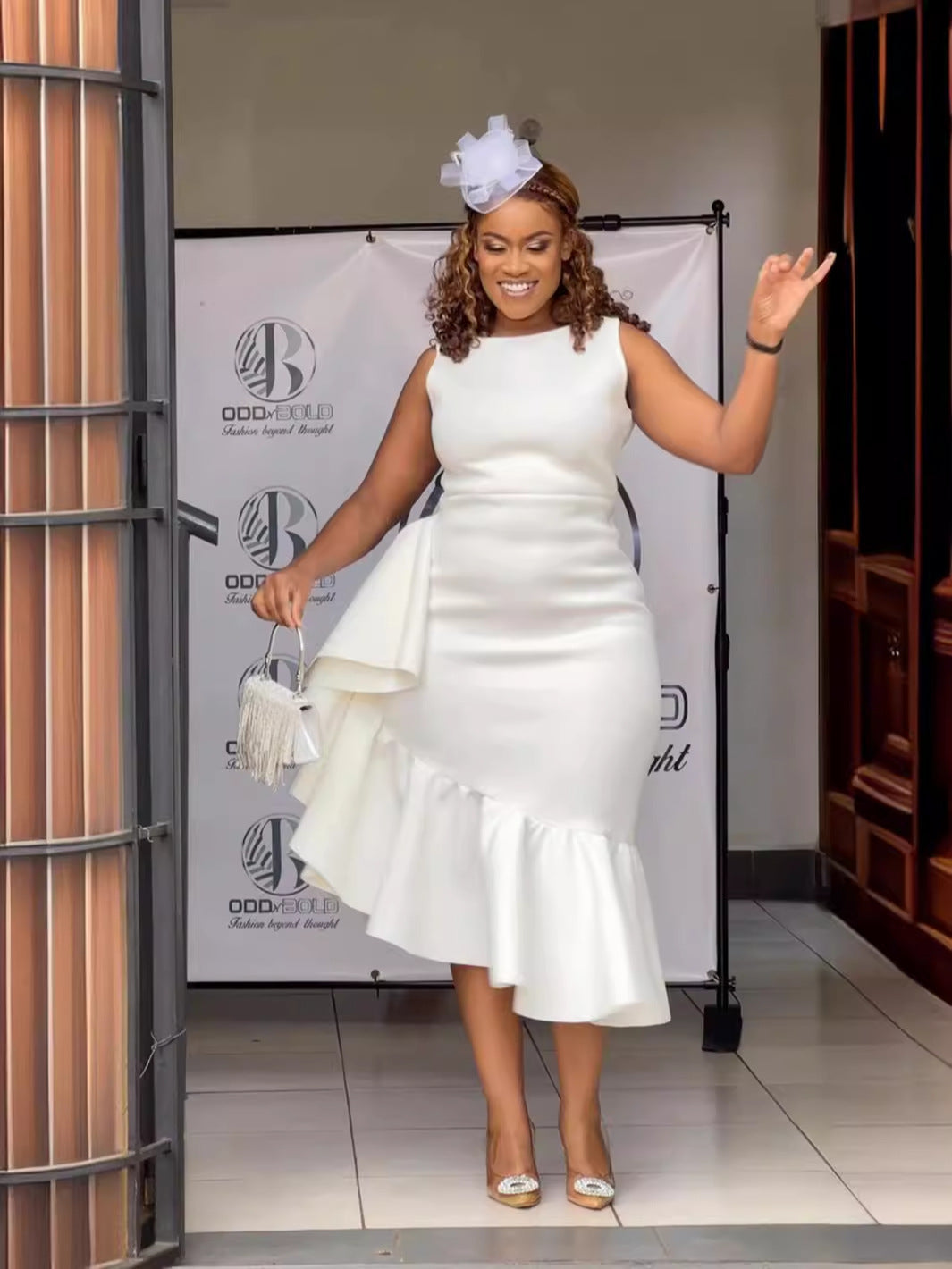 Woman wearing a white sleeveless dress with ruffle detailing and an asymmetrical hemline, accessorized with a white fascinator and silver handbag.